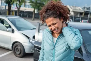 A woman gripping her neck with an automobile accident in the background